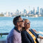 Two people smiling on a boat with a city skyline and water visible in the background under a clear sky.