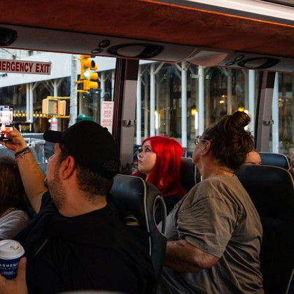People inside a bus, some taking photos through the windows. An emergency exit sign is visible, with a city street outside.