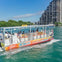A water taxi with passengers cruises on a greenish-blue body of water, with a modern high-rise building and trees in the background.