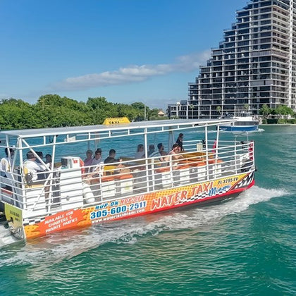 A water taxi with passengers cruises on a greenish-blue body of water, with a modern high-rise building and trees in the background.
