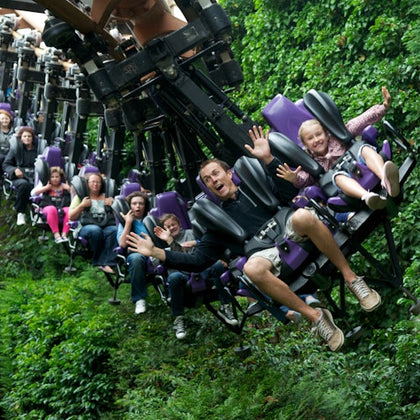 People on a rollercoaster with purple seats, surrounded by dense green foliage, some with hands raised and excited expressions.