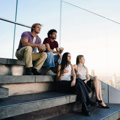 Four people sitting on stone steps holding drinks, with a glass railing and cityscape in the background.