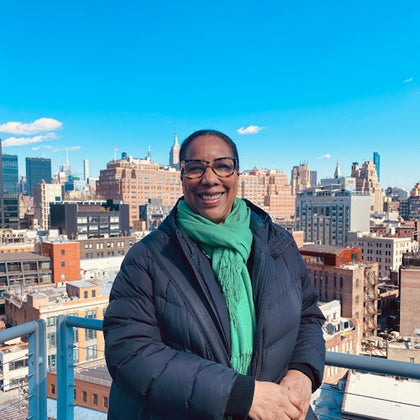 A person wearing glasses, a green scarf, and a dark coat stands on a balcony with a cityscape and blue sky behind them.