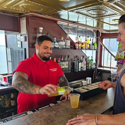 A bartender in a red shirt is pouring a drink for a customer at a bar with bottles in the background, on a boat.
