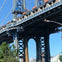 A view of a large suspension bridge structure with visible steel beams and a cityscape in the background under a blue sky.