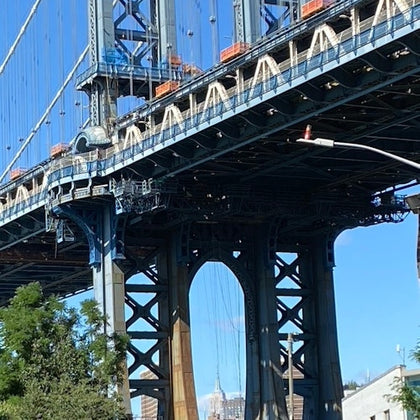 A view of a large suspension bridge structure with visible steel beams and a cityscape in the background under a blue sky.