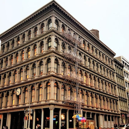 A historic multi-story building with arched windows, a fire escape, and a clock on the corner, set against a cloudy sky.