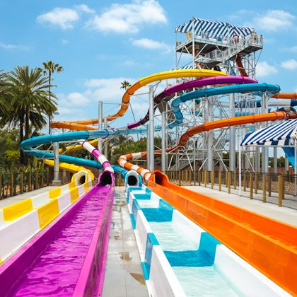 Colorful water slides and a tall slide tower in a water park under a sunny sky, surrounded by palm trees.
