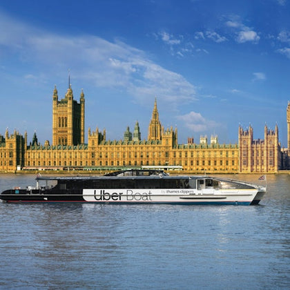An Uber Boat by Thames Clippers ferry on the Thames River in front of the Palace of Westminster under a blue sky.