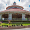 A large carousel housed in an ornate pavilion with a red roof, surrounded by landscaped greenery and flowers.