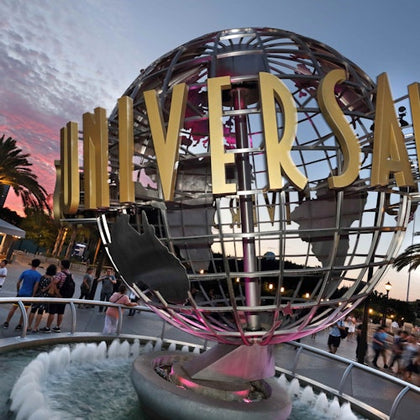 A large metallic globe with "Universal" written on it, surrounded by a fountain and people, during sunset with palm trees.