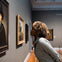 A person closely observing a framed painting in an art gallery, surrounded by other paintings on the walls.