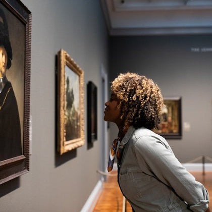 A person closely observing a framed painting in an art gallery, surrounded by other paintings on the walls.