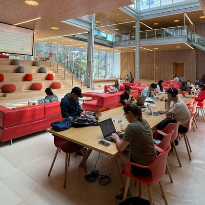People working on laptops at long tables in a modern, open space with red couches, wooden accents, and large windows.