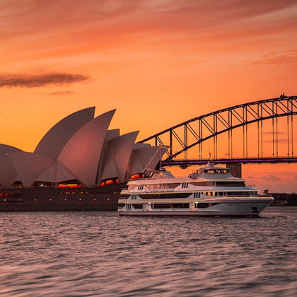 A cruise ship in front of the Sydney Opera House and Harbor Bridge during a vibrant orange sunset.