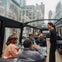 A group of people dining inside a glass-roofed bus with a waiter attending to them; city buildings are visible in the background.