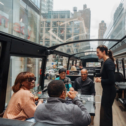 A group of people dining inside a glass-roofed bus with a waiter attending to them; city buildings are visible in the background.