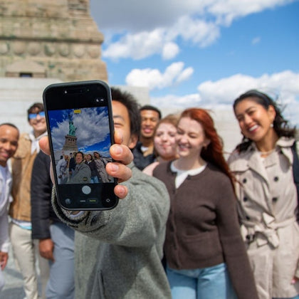 A group of people taking a selfie in front of the Statue of Liberty on a sunny day, with the phone showing the captured photo.