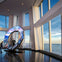 Tall windows showcase a skyline and ocean view. An exhibit with screens and "One World Trade Center" sign stands indoors with two people nearby.