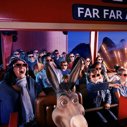 People wearing 3D glasses enjoy an indoor ride with a donkey character in front, background features mountains and lights. "FAR FAR" sign visible.