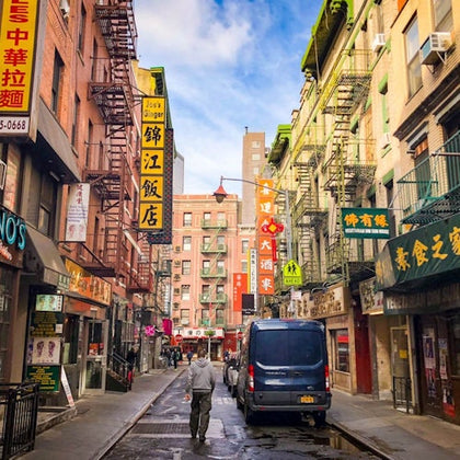A narrow street in Chinatown with colorful signs, fire escapes, shops, a blue van, and a person walking away from the camera.