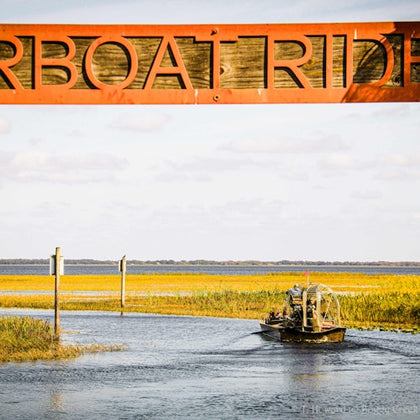 An airboat navigates a waterway surrounded by marshland, beneath a wooden "Airboat Ride" sign.