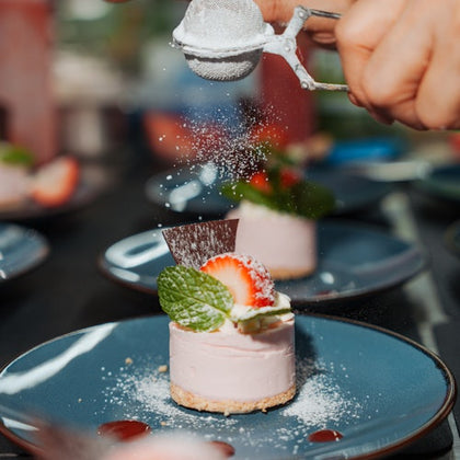 A hand dusts powdered sugar onto a strawberry cheesecake garnished with mint leaves on a blue plate.