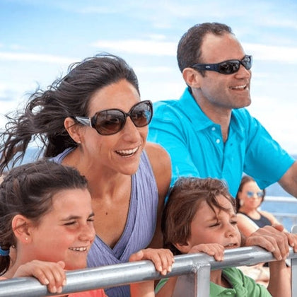 A family smiling and leaning on a railing outdoors, with bright sunlight and a blue sky in the background.