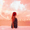 A person with red hair stands in a room filled with white balloons, wearing a patterned dress, under soft lighting.