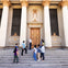 A group of people standing on the steps in front of a large building with columns and a decorative door.