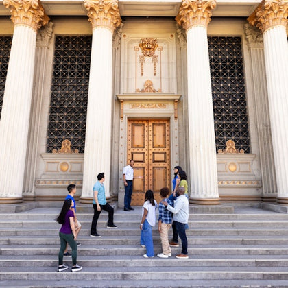 A group of people standing on the steps in front of a large building with columns and a decorative door.