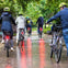 A group of cyclists wearing helmets ride along a wet, tree-lined path with tail lights visible.