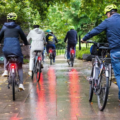 A group of cyclists wearing helmets ride along a wet, tree-lined path with tail lights visible.