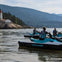 Three people on jet skis near a rocky shoreline with a lighthouse and building, under a clear sky with mountains in the background.