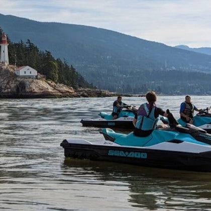Three people on jet skis near a rocky shoreline with a lighthouse and building, under a clear sky with mountains in the background.