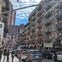 A busy urban street with pedestrians, cars, and buildings adorned with hanging decorations under a partly cloudy sky.