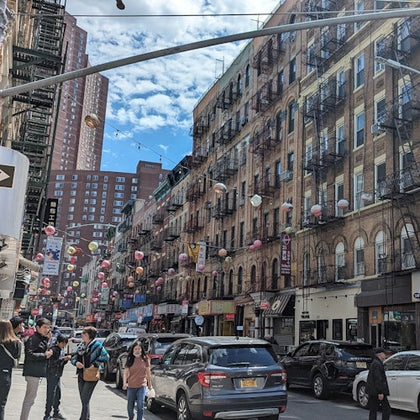 A busy urban street with pedestrians, cars, and buildings adorned with hanging decorations under a partly cloudy sky.