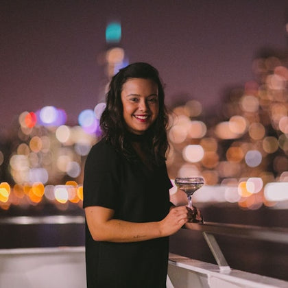 A woman in a black dress holding a glass stands on a balcony at night, with city lights blurred in the background.