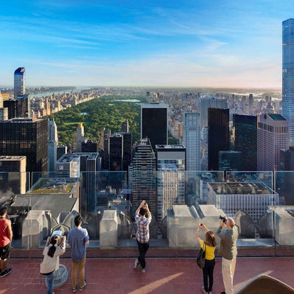 People look out over a cityscape with tall buildings and a large green park, likely Central Park, on a sunny day.
