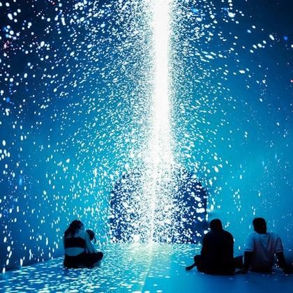Three people sit on the floor in a dark space illuminated by cascading white lights against a blue background.