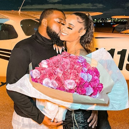 A man kissing a woman who is holding a large bouquet of pink and purple roses, with a small airplane in the background.