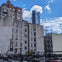 A cityscape featuring mid-rise buildings, a parking lot with cars, and a tall skyscraper in the background under a blue sky.
