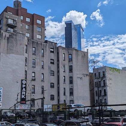 A cityscape featuring mid-rise buildings, a parking lot with cars, and a tall skyscraper in the background under a blue sky.