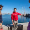 A person in a red hoodie gestures near a waterfront with moored sailboats under a clear blue sky, with others nearby.