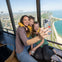 A woman and two children sit near a window of a tall building, smiling and taking a selfie with a scenic city and lake view.