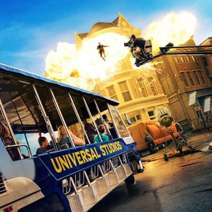 People on a Universal Studios tour bus watch a movie scene being filmed with explosions and a stunt person flying through the air.