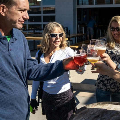 Four people clinking glasses of beer in a casual outdoor setting during daylight.