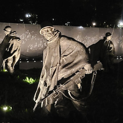 Statues of soldiers holding rifles in a nighttime memorial, with a reflective wall in the background.
