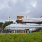 A space shuttle mounted on top of a Boeing 747 with a building and vegetation in the foreground under a cloudy sky.