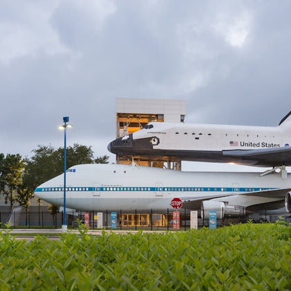 A space shuttle mounted on top of a Boeing 747 with a building and vegetation in the foreground under a cloudy sky.